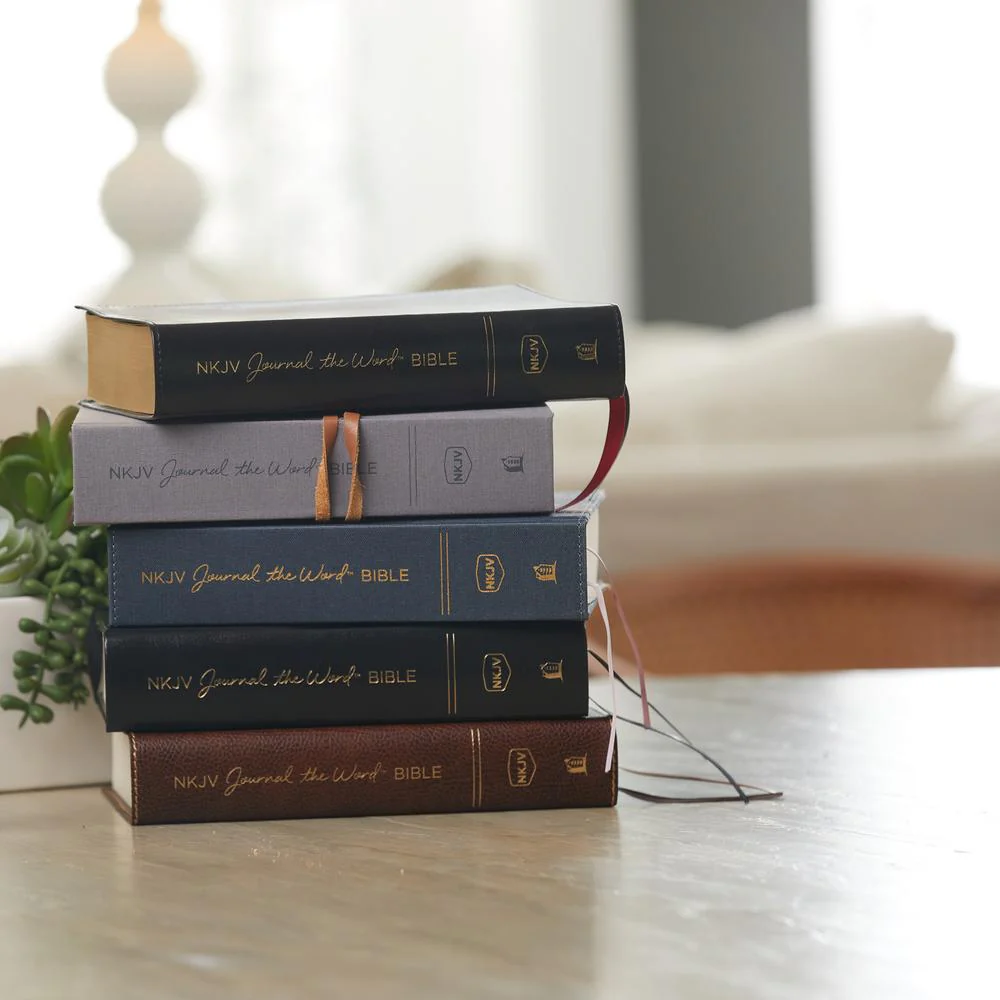Stack of Journal the Word Bible editions in various bindings and colors on a light colored table, showing available formats and premium craftsmanship.