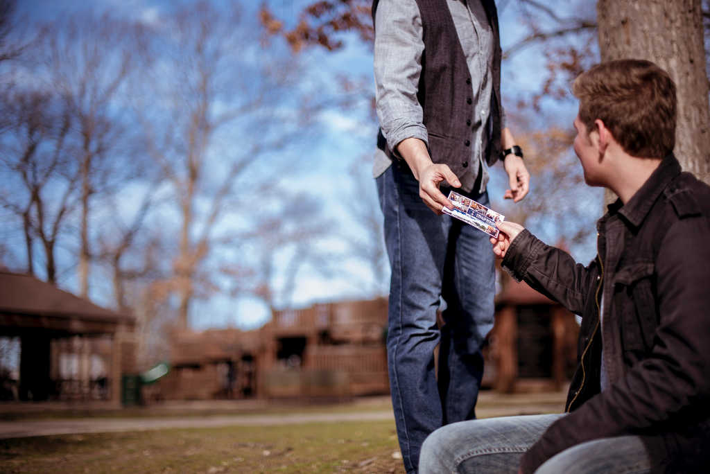man sitting outdoors handing church invitation to man walking by